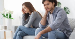 Couple sitting on a sofa, both with hands on their heads, looking stressed or worried in a bright living room.
