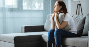 Woman sitting on a gray sofa, holding a pillow to her chest and gazing pensively out a window.
