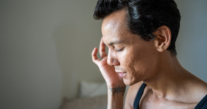 Profile of a shirtless man with short dark hair, eyes closed, touching his temple with one hand, a tattoo visible on his forearm.