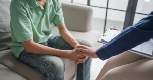 Therapy session: client in a green polo sits on a sofa while a counselor holds their hands in a supportive moment with a clipboard nearby.