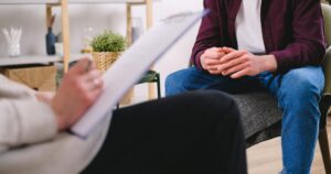 Therapist takes notes on a clipboard while a client sits with hands folded in a cozy office setting.