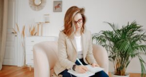 Woman with red glasses writing in a notebook while seated on a pale pink sofa at home.