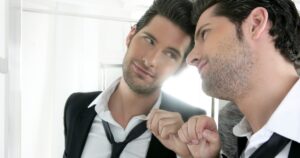 Two men in business attire share a moment in a bright room, one gently tightening the other's loosened tie while they smile at their reflection in a mirror.