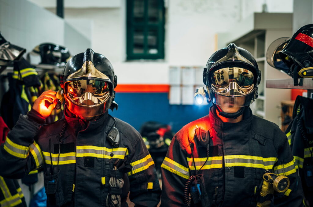 Two firefighters in full turnout gear stand in a fire station, wearing shiny helmets and reflective stripes on their jackets.