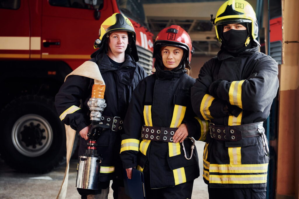 Three firefighters in turnout gear pose inside a fire station, with a red fire truck behind them.