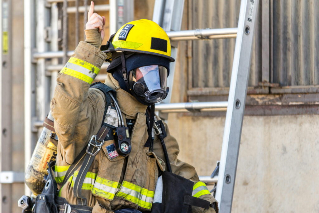 Firefighter in full turnout gear and a yellow helmet gives a thumbs up, beside a metal ladder on a construction site.