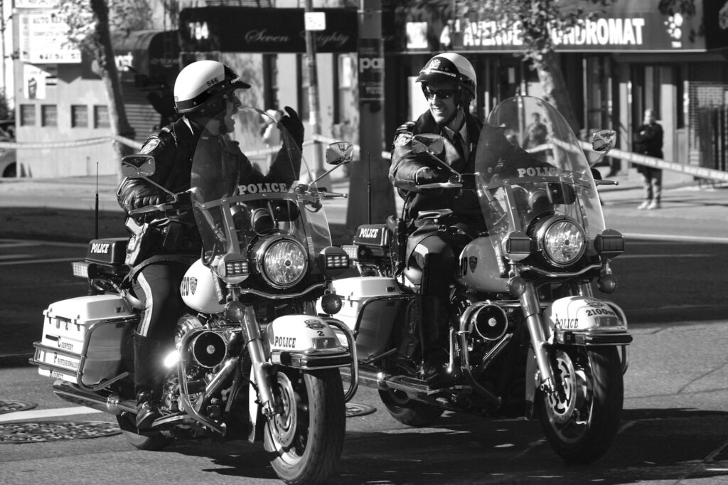 Two police motorcycles with riders in helmets stopped on a city street, storefronts in the background.”