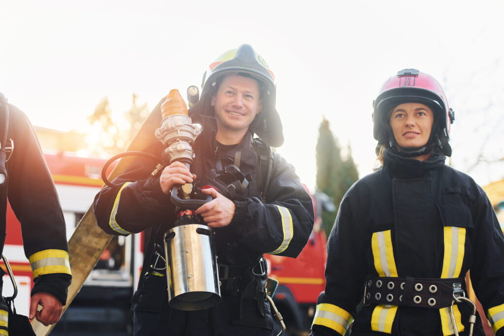 Two firefighters in full turnout gear, one holding a large hose nozzle, with a fire truck in the background.